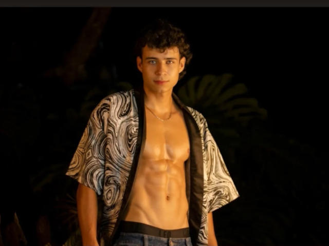 SofiaAndgeordi, with curly brown hair, wearing an open, patterned, short-sleeve shirt, exposing a toned, muscular, bare chest, and black waistband, standing against a dark background.