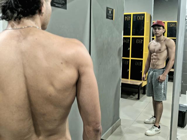 Los músculos de la espalda de SofiaAndgeordi son visibles, está sin camisa, con pantalones cortos grises y una gorra roja, y está de pie frente a un espejo que refleja su abdomen tonificado, musculoso y definido..