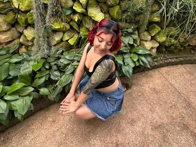 JuanaAhh mit rot gefärbten Haaren, schwarzem Crop-Top und Jeansrock, in die Hocke gegangen, zeigt tätowierten Arm, kleine Brüste und ein süßes Lächeln, in einem üppigen, grünen Garten..