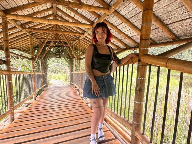 Juana, avec des cheveux roux, un haut en cuir noir, une jupe en jean, des tatouages sur les jambes, debout sur un pont en bambou, tenant la rambarde, souriante, portant des baskets blanches, sous un toit en bois..