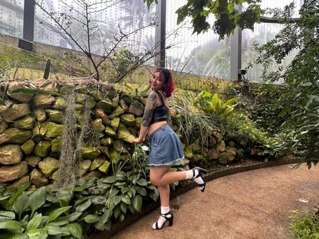 JuanaAhh, con el cabello teñido de rojo, brazos tatuados, un top corto negro, una falda vaquera y sandalias blancas, apoyada contra una pared de roca con agua, en un jardín verde y brumoso..