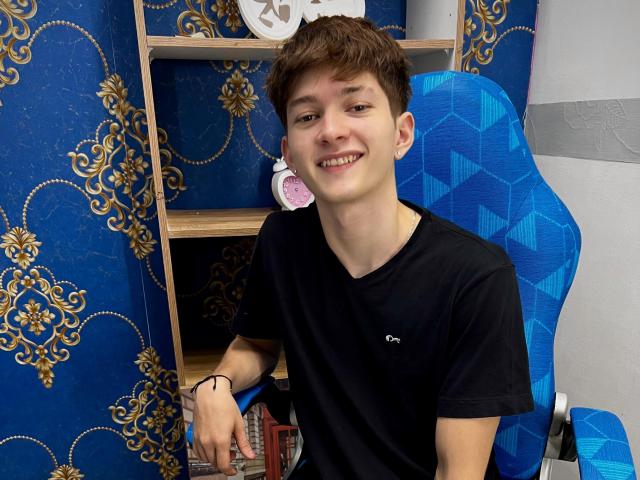 JakeDavins, young dude with short brown hair, smiling, wearing a black t-shirt, sitting on a blue patterned chair, holding a book, black wristband, casual, cute.