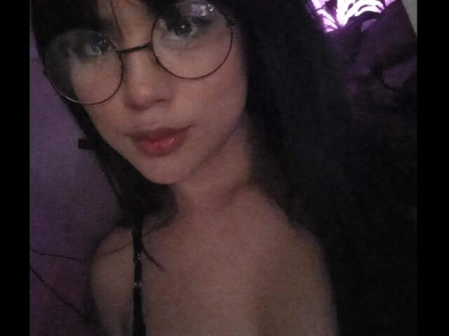 SteffanyTaylor with glasses, dark hair, and red lipstick, showing her bare shoulders and small tits, looking into the camera with a slight smile. Dimly lit, purple background.