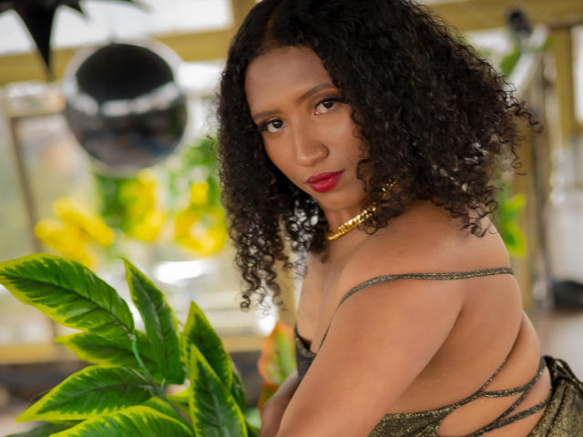 KarinaLovey with curly black hair, wearing a gold necklace and green strappy dress, looking over her shoulder, showing her back and red lips, with a green plant in front.