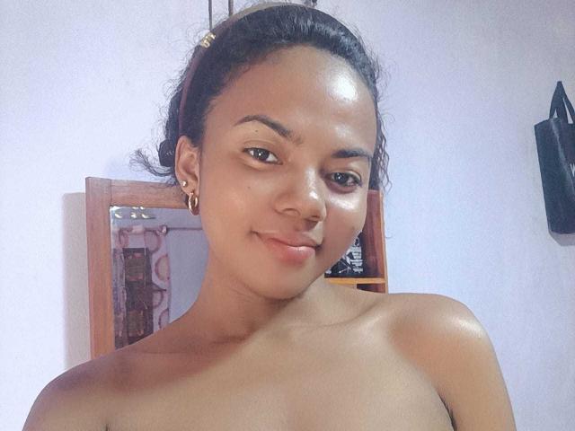 ElvaSexy has a cute smile, brown skin, and small earrings, with her dark hair in a bun, showing her bare shoulders and upper chest. She looks sexy and confident.