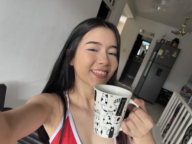 DeisyWest with long black hair, smiling, holding a white mug with black and yellow patterns, wearing a red and white top, in a kitchen. Her tits are small.