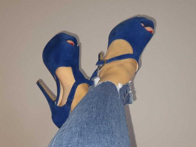 Le gambe di ElizabethRaegan, con le sue scarpe con il tacco alto blu e i jeans blu leggermente strappati, che mostrano le unghie dei piedi dipinte di rosso. Le sue caviglie sono incrociate, mettendo in mostra il suo calzature elegante e sexy..