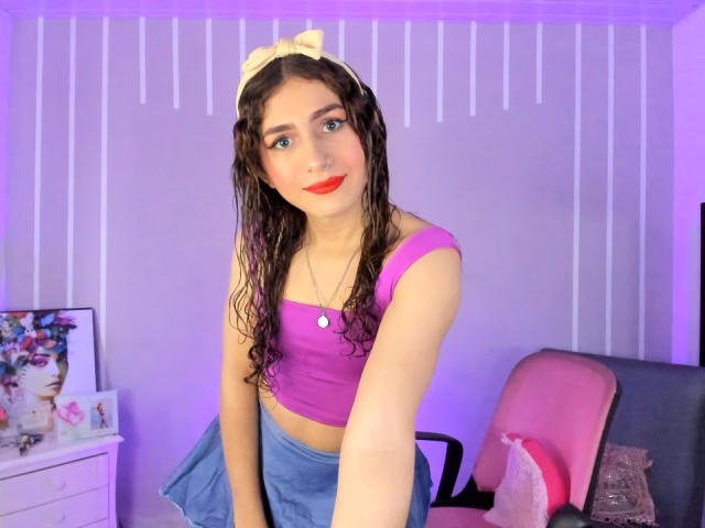 AndyDupont with wet brown hair, pink crop top, blue skirt, yellow bow, red lipstick, and a necklace, leaning forward, showing a cute smile, and small tits.