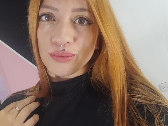 BlondeRouss, avec des cheveux roux et orange, un piercing au nez et des lèvres roses, portant un haut noir, des yeux marron, un eye-liner noir et une bague à la main gauche..
