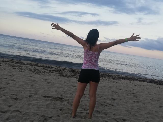 LunaVenus stands on a beach with arms outstretched, wearing a pink patterned top and black shorts, facing a calm ocean at sunset. Her ass is visible.
