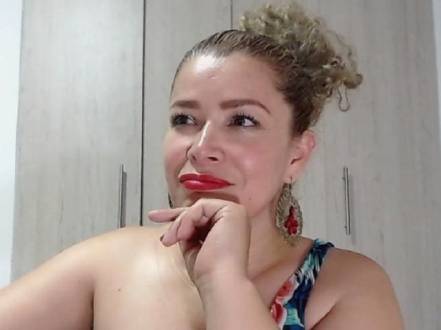 DalilaSuperHot with curly blonde hair, red lipstick, and red earrings, resting her chin on her hand, smiling, showing her bare shoulders, wearing a colorful, patterned top.