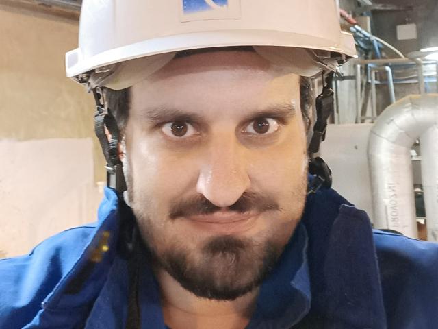 SexyJS portant un casque de chantier blanc, une chemise bleue et une barbe, souriant avec des yeux marron, des cheveux foncés légèrement bouclés et une petite moustache. Son nez est un peu plat..
