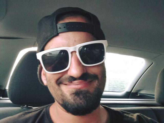 SexyJS indossa un berretto nero da baseball e occhiali da sole bianchi oversize, con un baffo e una barba, e sorride seduto in un'auto. Si vede la sua camicia marrone..