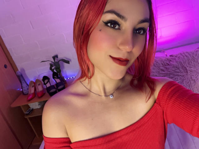 AmyStreetwise69 with red hair, off-shoulder red top, black eyeliner, nose piercing, necklace, smiling, white skin, and pink-purple lighting, showing her tits and ass in the selfie.
