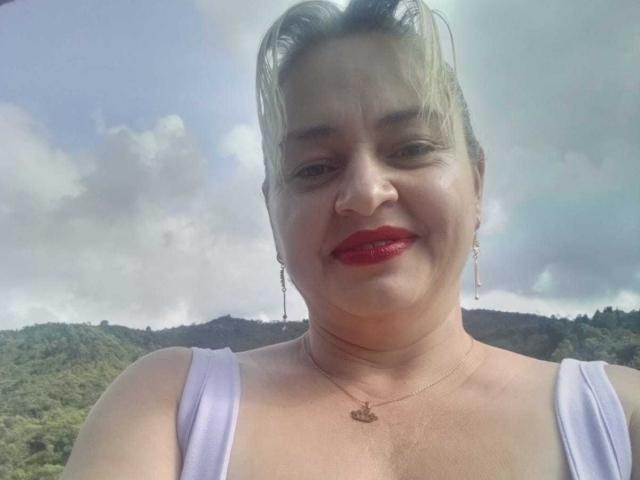 SalomeAriza with red lipstick, smiling, wearing a white tank top, necklace, and earrings, has blonde hair in a messy updo, and a slightly chubby face with light skin.