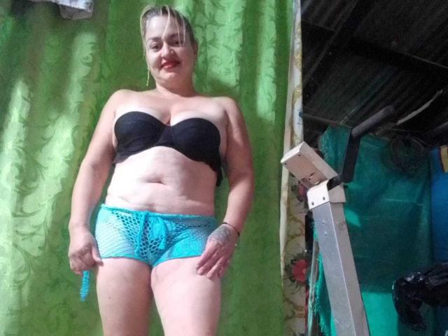 SalomeAriza, blonde, smiling, wearing black strapless bra, showing her tits, and turquoise fishnet shorts, standing with hands on her thighs, inside a cluttered, green-walled room.