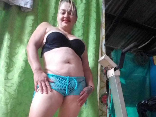 SalomeAriza with blonde hair, black strapless bra, and blue fishnet shorts, standing, smiling, hand on hip, visible tattoo, indoor setting, low angle shot.