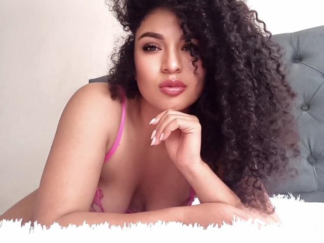 CarlaMuille with curly black hair, pink bra, and pouty lips, resting her chin on her hand, showing her tits and pink lingerie, lying on a white fluffy surface.