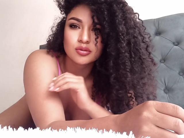 CarlaMuille, con i capelli neri ricci, un reggiseno rosa e labbra piene, sdraiata su un letto, mani vicino al viso, che guarda la fotocamera, sexy, carina, ambientazione: camera da letto..