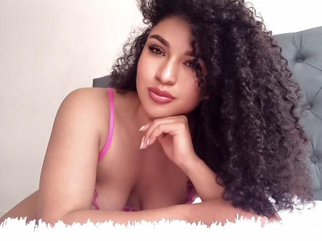 CarlaMuille with curly black hair, pink bra, and subtle makeup, resting her chin on her hand, showing her medium-sized tits, with a soft smile and long nails.