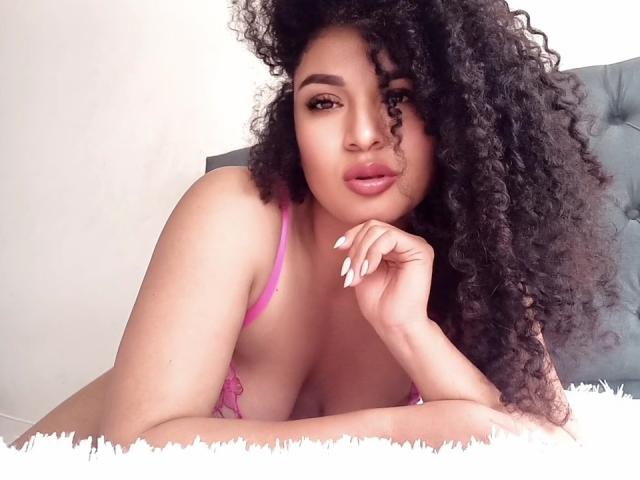 CarlaMuille with curly black hair, pink bra, and white nails, resting her chin on her hand, showing her tits, with a sultry look, lying on a fluffy white surface.