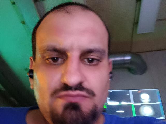 SexyJS a une tête chauve, des sourcils foncés, une moustache et une barbe de chèvre. Il porte des écouteurs noirs et une chemise bleue, avec une lumière verdâtre sur le côté gauche..