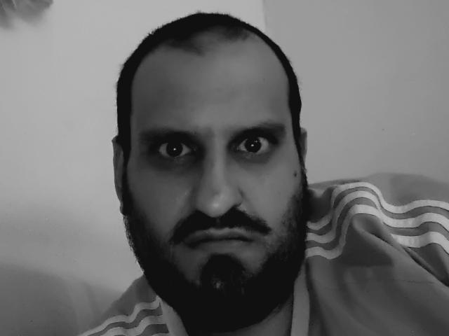 SexyJS a une expression sérieuse, une barbe noire et une moustache avec un bouc. Il est chauve, avec des sourcils foncés, et porte une chemise rayée. Photo en noir et blanc..