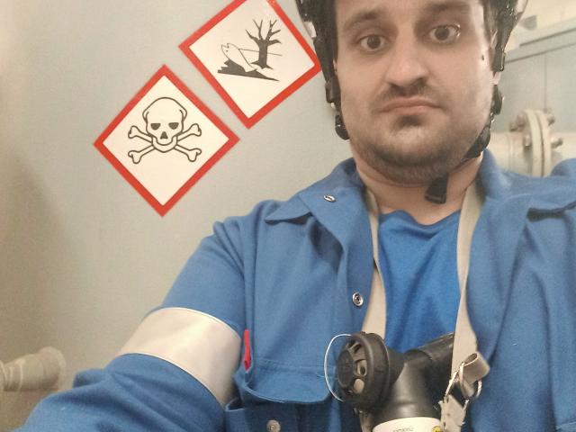 SexyJS met een helm, een blauw shirt en een witte armband, die een zwart pistool vasthoudt, een headset draagt en een serieuze uitdrukking heeft, met schedel- en botenkunst aan de muur..