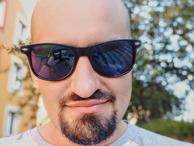 SexyJS a une tête chauve, des lunettes de soleil noires et une petite barbe de style "chou", et porte une chemise grise avec un motif discret. On voit des reflets dans les lunettes, et il a un léger sourire. La scène se déroule en extérieur..