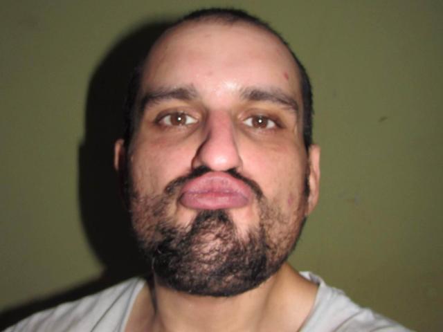 SexyJS ha la testa calva, gli occhi marroni e una barba folta e riccia. Sta facendo una smorfia, con le labbra leggermente strette e di un rosa sporco. Indossa una camicia bianca..
