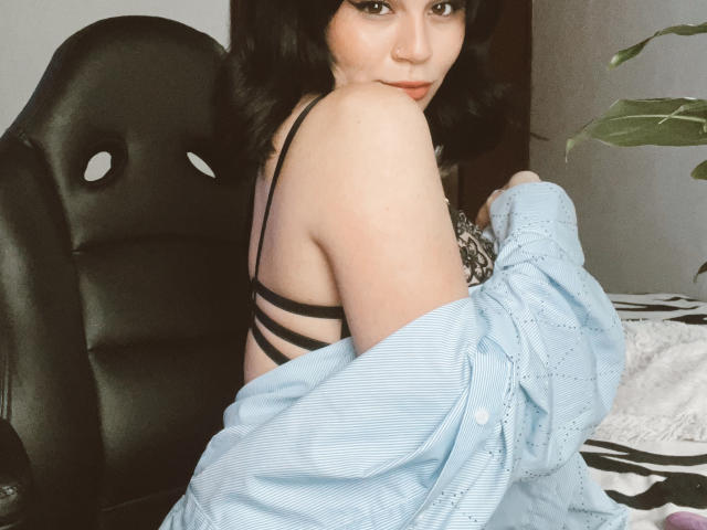 ShantallParis with black hair, light skin, and small tits, wearing a black strappy bra, light blue shirt off, looking back, sitting in a black chair, and potted plant nearby.