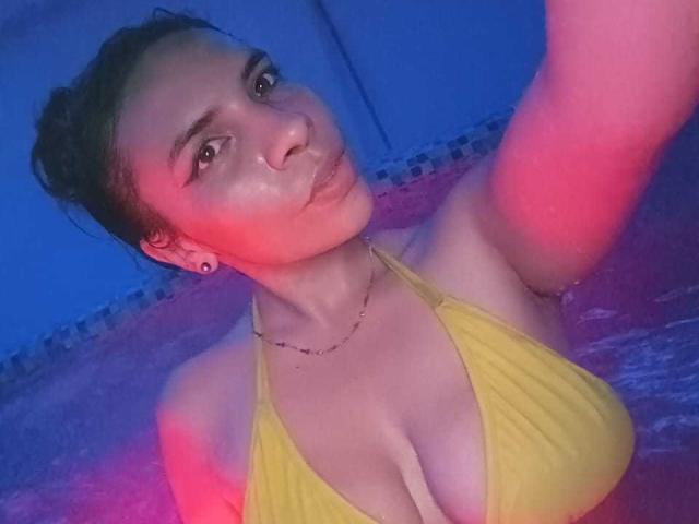 VioletaStone with pouty lips, wearing a yellow bikini, showing big tits, in a blue and pink lit pool, arm raised, dark hair in a bun, small earrings.