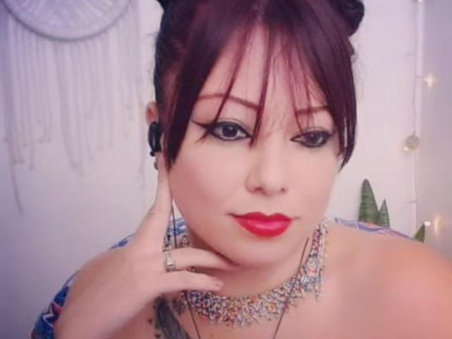 Ccristall has red lipstick, black eyeliner, and a jeweled necklace. She's touching her chin with a hand showing a ring. She has red hair in a bun.