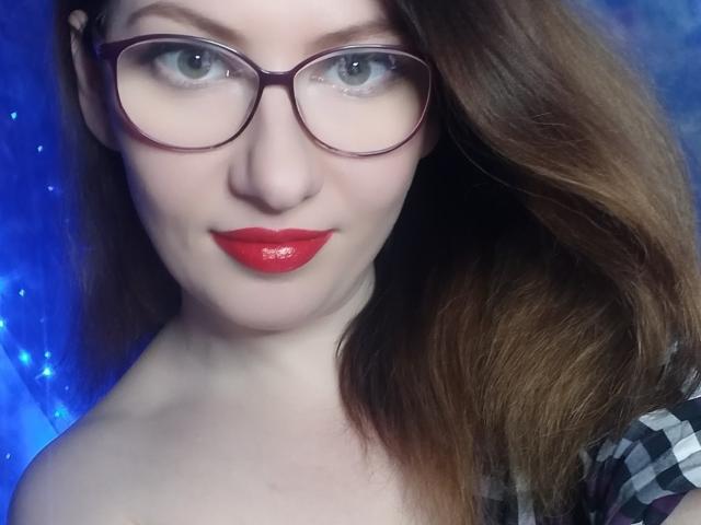 PaulinaSunny has red lipstick, glasses, and brown wavy hair, with fair skin and blue eyes, showing off her tits in a selfie with a blue background.