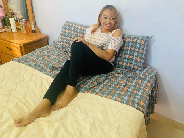 AralySex-hot sits on a bed with a floral-patterned cover, wearing a pink off-shoulder top and black pants, barefoot, legs crossed, smiling, in a bedroom.