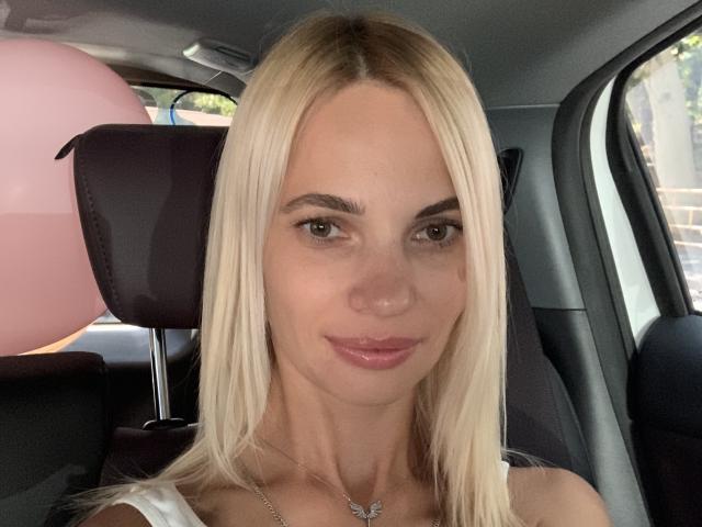 DonorDi has blonde hair, green eyes, and glossy pink lips, wearing a white top with a silver necklace, sitting in a car with a pink balloon behind her.
