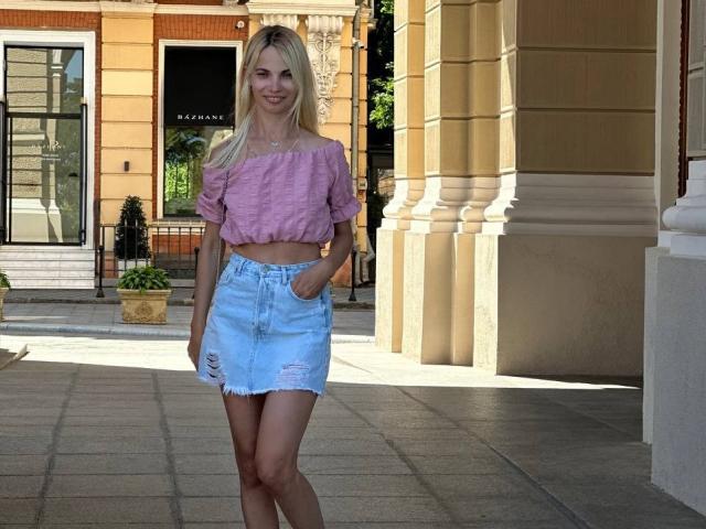 DonorDi with blonde hair, pink off-shoulder crop top, and blue distressed denim skirt, standing in front of a beige building, smiling with hands in pockets.
