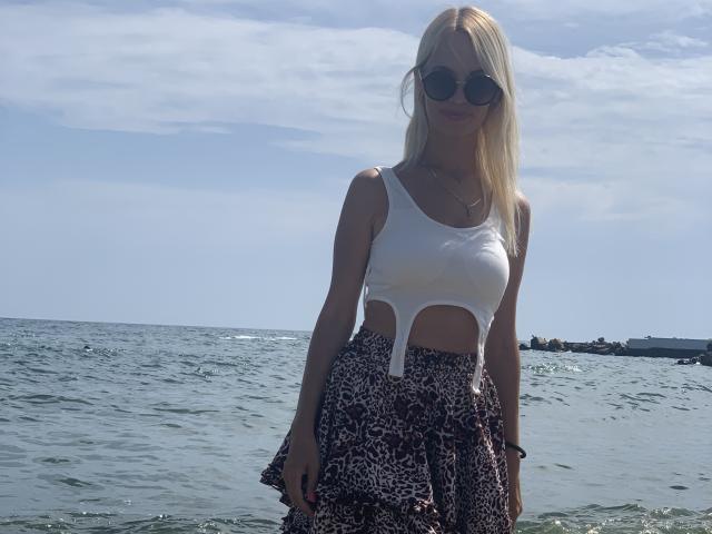 DonorDi with blonde hair, wearing round sunglasses, white cutout top, and leopard print skirt, standing in shallow ocean water. Boobs visible through top.