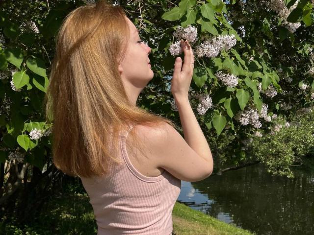 Radiants: Una persona con cabello rojo, camiseta rosa, sosteniendo lilas blancos, mirando hacia arriba, de pie cerca de un árbol verde, junto a una superficie de agua reflectante, en un día soleado..