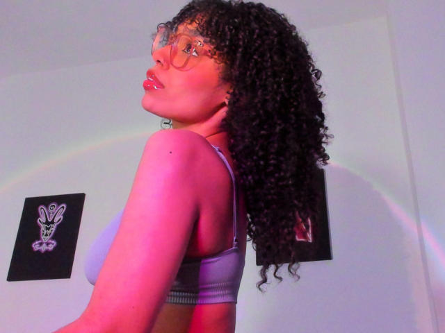 NickiBloom with curly black hair, glasses, and purple sports bra, showing her tits, pink lips, and a subtle rainbow reflection in the background.