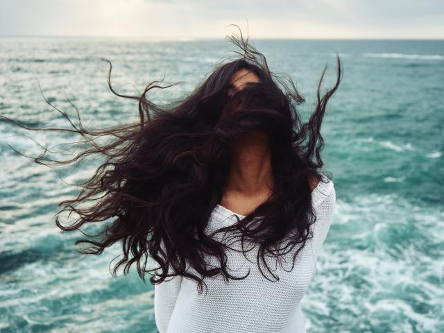 YaraTully with long, wild, dark brown hair covering her face, wearing a white, long-sleeved, textured shirt, standing in front of a green, choppy ocean.