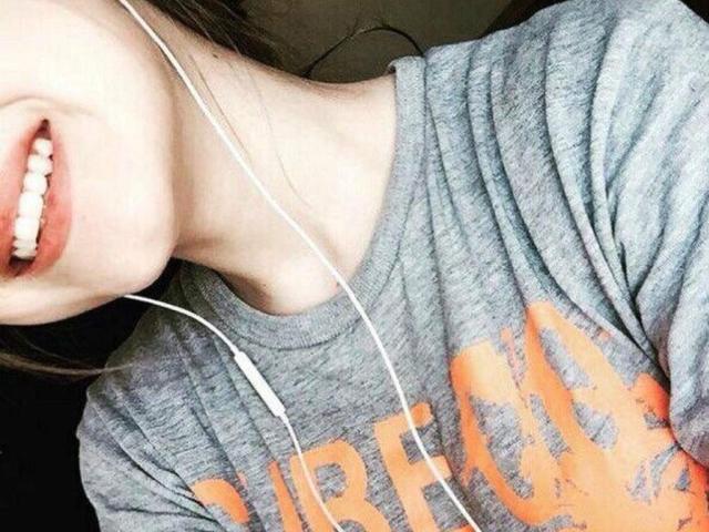 YaraTully's face with a smile, wearing a gray shirt, white earbuds in, showing her teeth and part of her neck, her lips slightly parted.