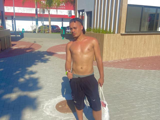 Shirtless Gabrielzao with tan skin, black shorts, and a black cap, holding a white bag, standing on a sunny sidewalk, shadow on his right, small brown circular metal cover.