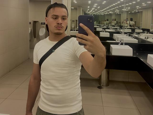 Gabrielzao with a white tee, black crossbody strap, and grey pants, taking a mirror selfie in a bathroom, holding a blue phone, with a serious expression.