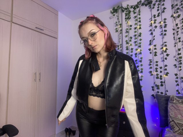 SarahRousen with pink hair, glasses, black lace bra, black leather jacket, white sleeves, black leather pants, standing in room, looking down, slightly bent forward, plant wall decor.