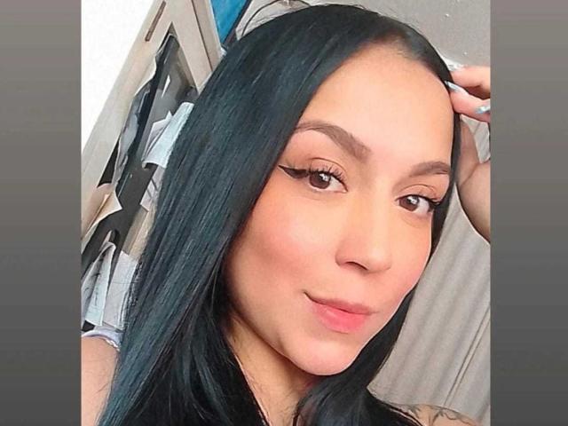 MissLitt has long black hair, brown eyes, and is smiling with slightly parted pink lips. She's touching her hair with one hand, showing off her winged eyeliner.