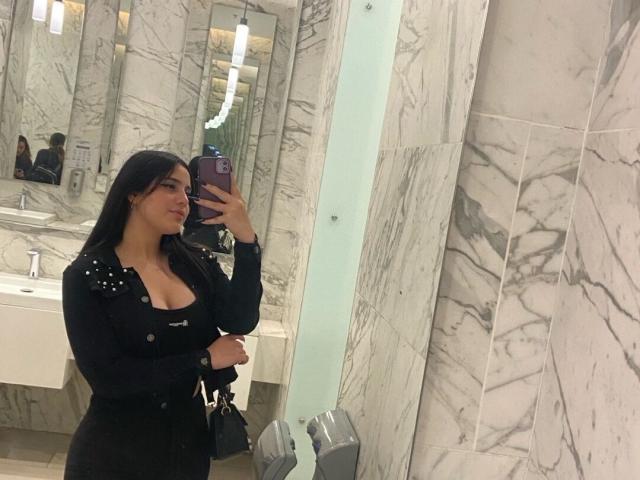 ChloePrie has large tits, wearing a black shirt with studs, taking a mirror selfie in a marble bathroom, holding a purple phone, smiling, long black hair, white sink behind.