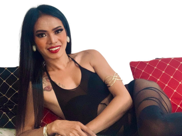 MistressMargaTS has long black hair, red lipstick, and is wearing a black see-through dress with a gold armband, showing small tits and black stockings, smiling at the camera.
