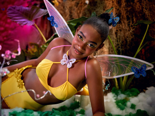 DalilaQueen met donkere huid, een geel bikinnetje, vlinderhaarspeldjes, feeënflappen, lachend, roze oogschaduw, een zichtbare tepel, een kleine vlindersketting, liggend op groen en wit, pluizig "gras".".