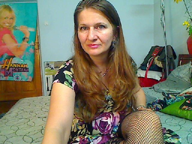 A DelightMature tem cabelos castanhos longos, batom rosa e veste um vestido floral com meias de rede, sentada em uma cama com um travesseiro verde. Seus seios estão visíveis..